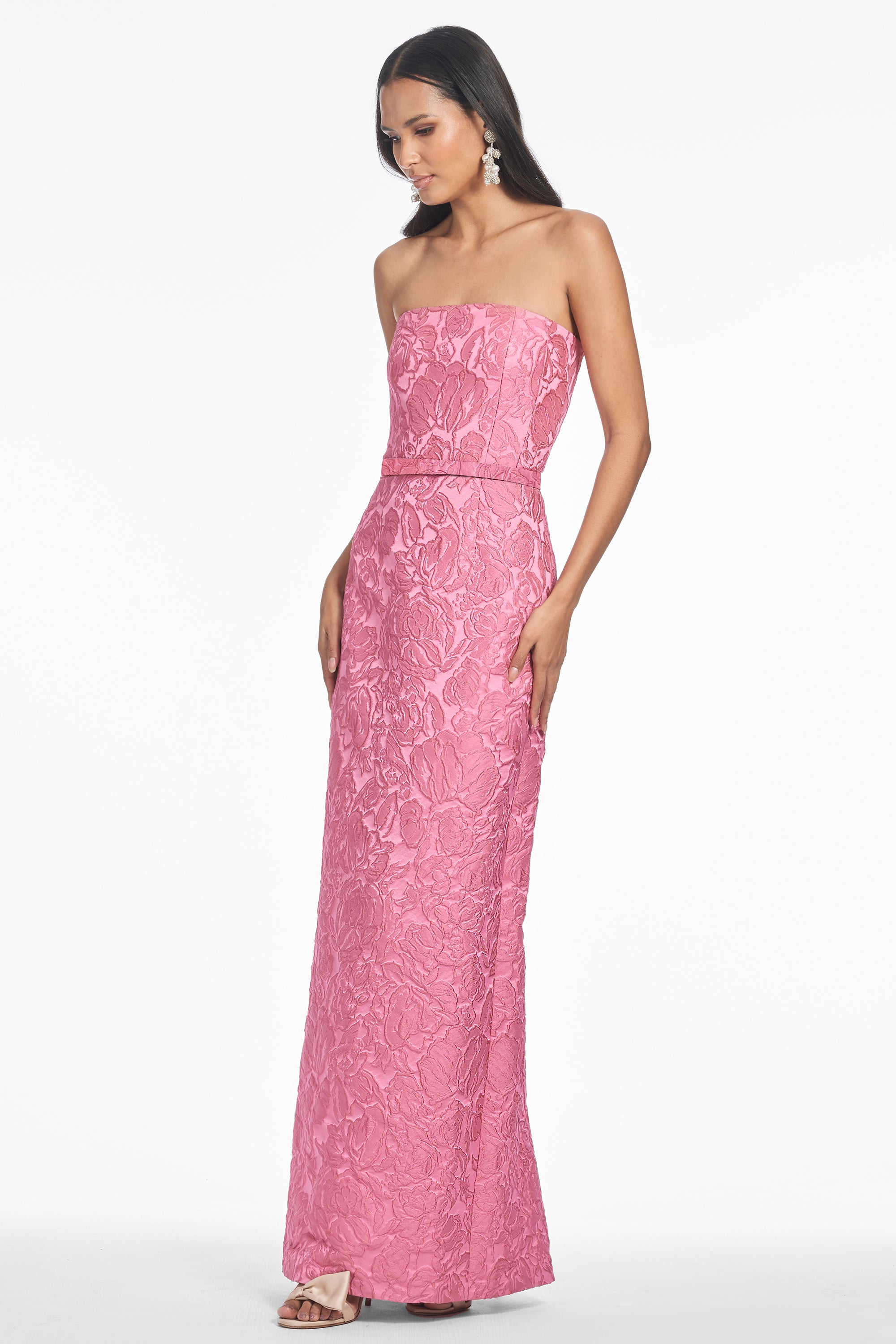 A woman wearing a rose pink, strapless, full-length gown with a column silhouette. The gown features a back zipper, full lining, back vent, and an attached belt.