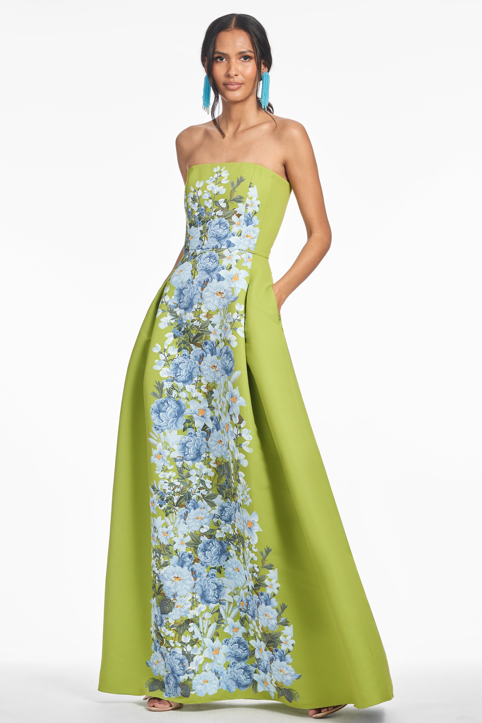 A woman wearing a strapless, peridot green, floral print gown with a fit and flare silhouette, featuring a cascade of flowers on the front.