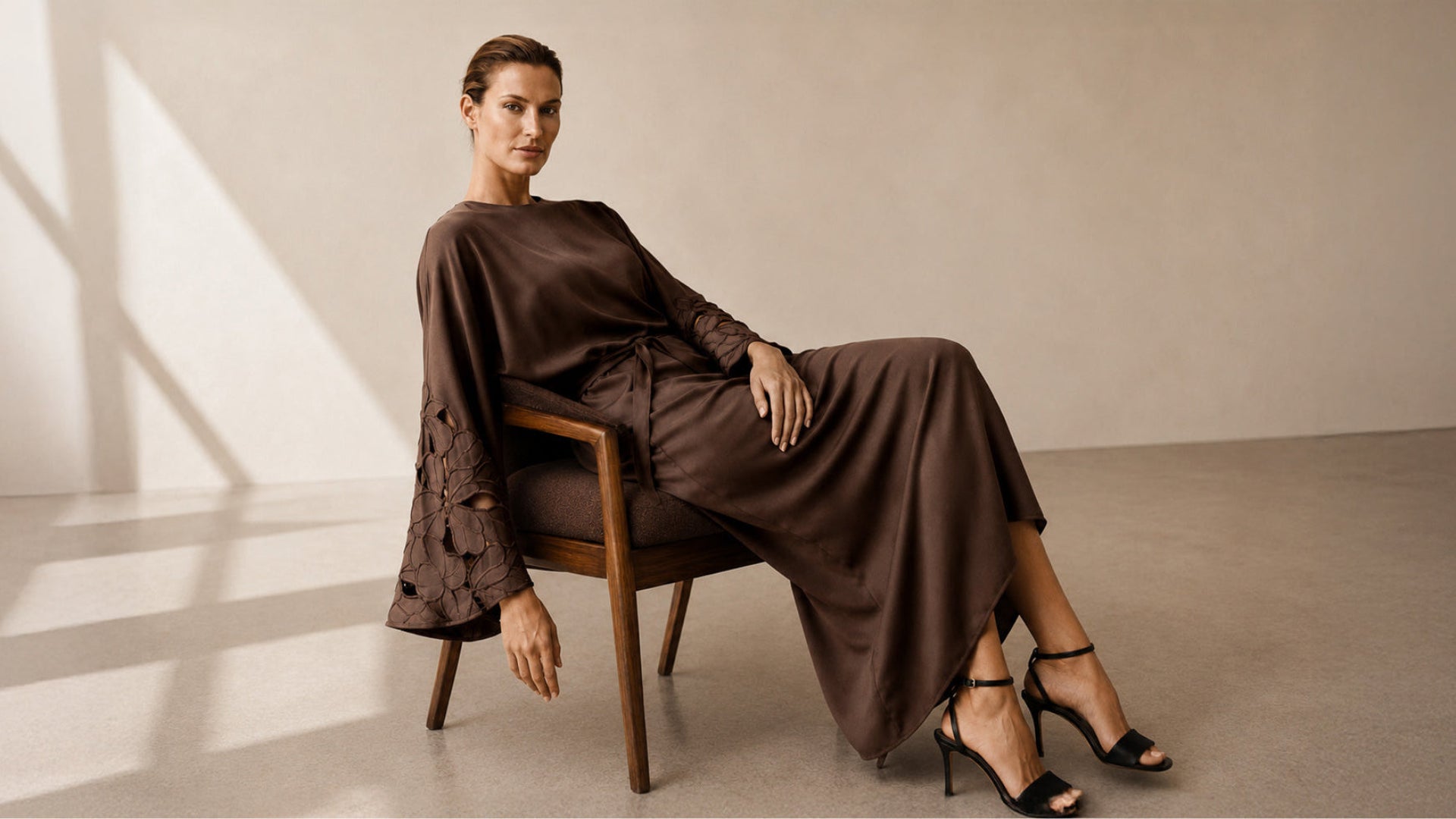 A woman seated on a wooden chair wearing a long sleeve brown satin like dress with lace detailing on one sleeve, posed in a minimalist studio with warm beige walls and floor