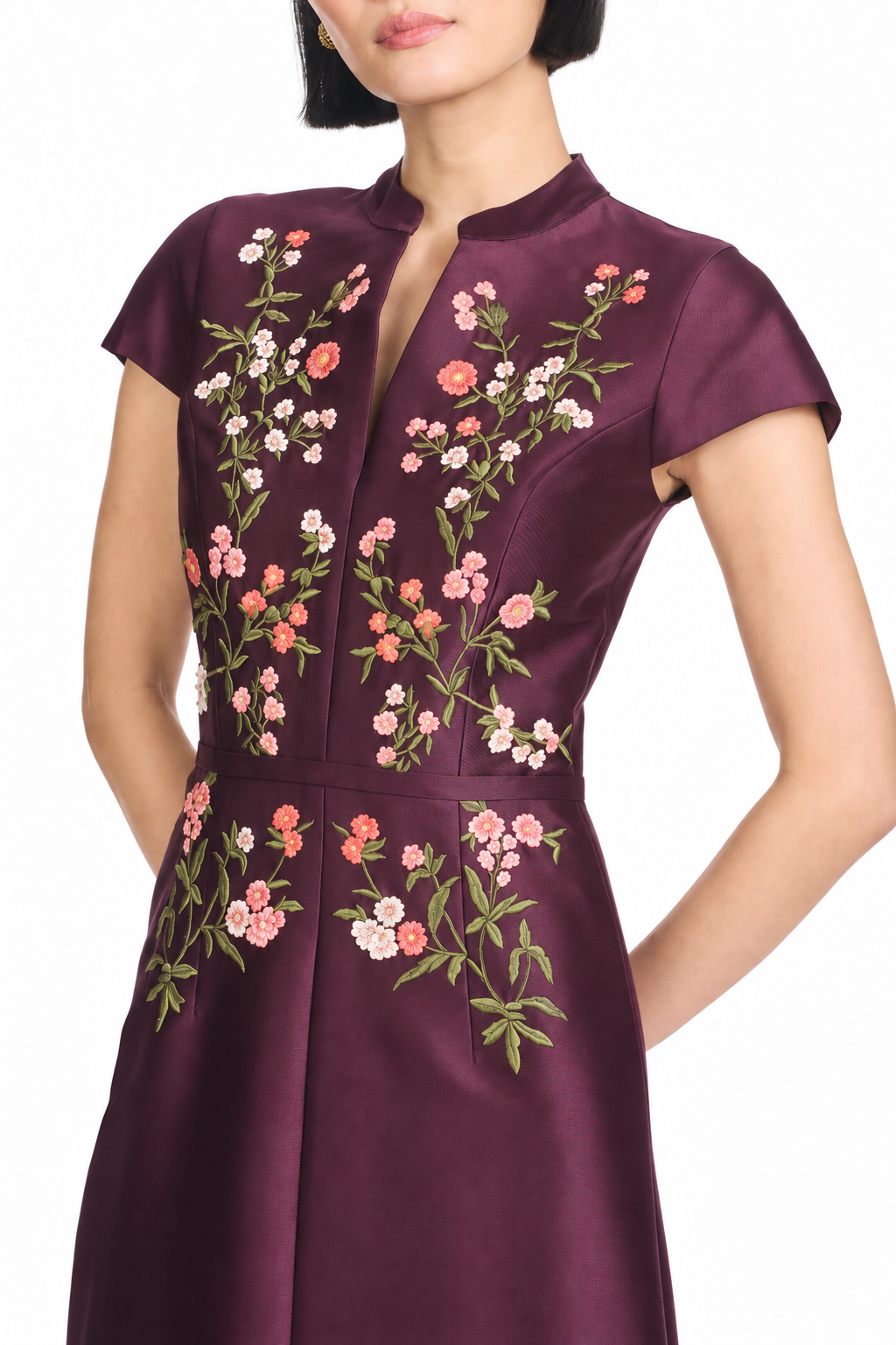A woman wearing a short-sleeve, high-neck dress in deep plum purple with detailed floral embroidery in pink, peach, and green on the bodice and skirt, shown against a plain white background.
